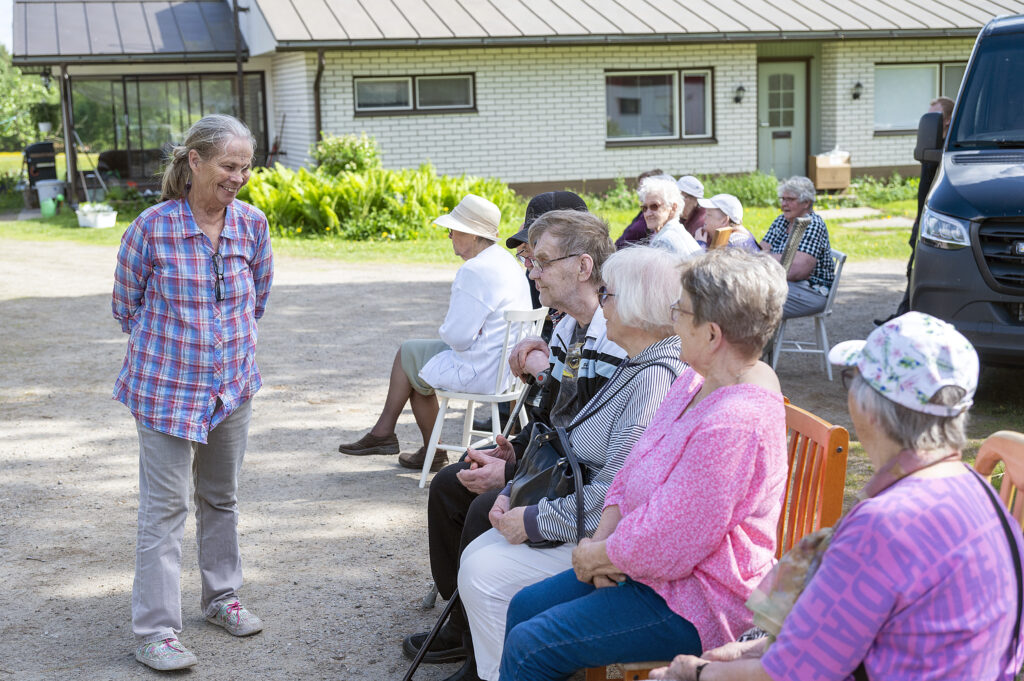 Nainen seisoo seniorikerhon jäsenten edessä ja katsoo heitä hymyillen.