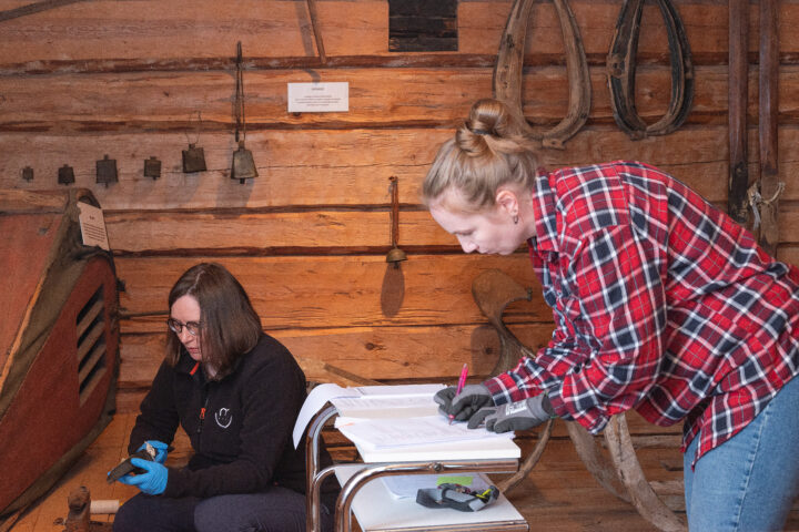 Museon työntekijä kirjaa muistiin huomioita esinekokoelmasta. Amanuenssi on kyykistynyt lattialle ja pitelee esinettä kädessään.