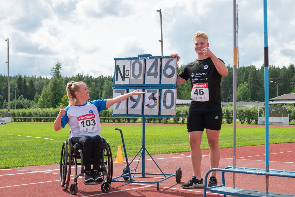 Keihäänheittäjät Jasmine Repo ja Jere Murto poseeraavat tulostaulun edessä. Tulostaululla on Murron juuri heittämä tulos 79,30. Molemmat näyttävät peukaloa ja Repo ojentaa kättään esitelläkseen Murron tulosta.