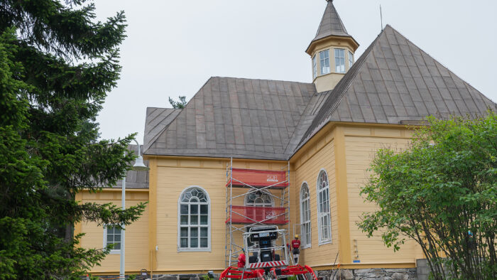 Pihtiputaan kirkko, jonka edustalla on maalausurakoitsijan teline, nosturi ja ikkunanpieliä maalaava työntekijä.