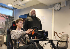 Kaksi henkilöä vierekkäin. Etualalla nuorehko henkilö pitelee käsissään rattia ja istuu. Toinen henkilö seisoo ja katsoo samaan suuntaan ajajan kanssa.