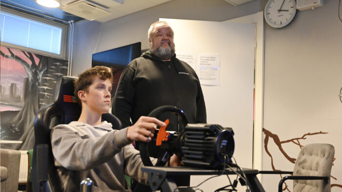 Kaksi henkilöä vierekkäin. Etualalla nuorehko henkilö pitelee käsissään rattia ja istuu. Toinen henkilö seisoo ja katsoo samaan suuntaan ajajan kanssa.