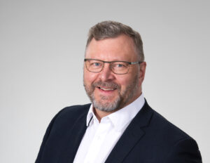 Mieshenkilö jolla silmälasit, musta takki ja valkoinen kauluspaita. Puolilähikuvassa.