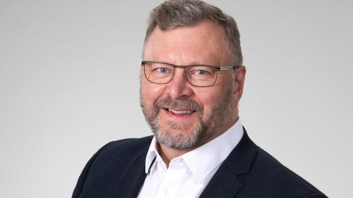 Mieshenkilö jolla silmälasit, musta takki ja valkoinen kauluspaita. Puolilähikuvassa.