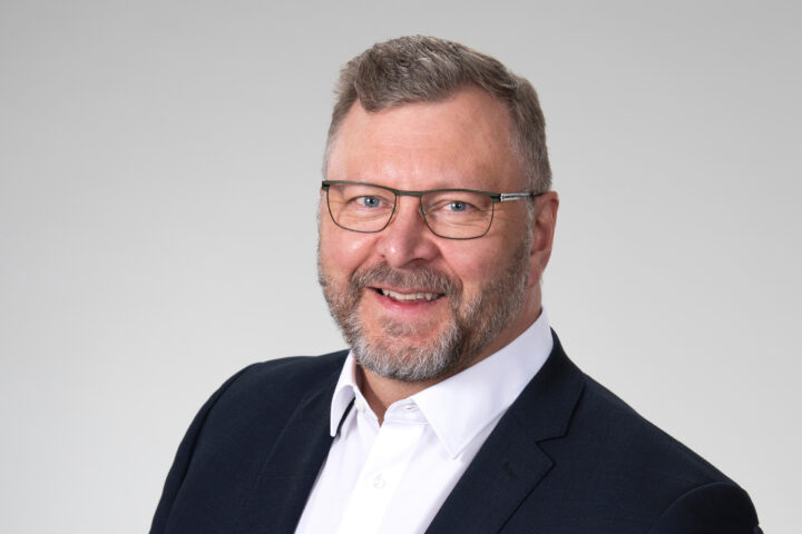 Mieshenkilö jolla silmälasit, musta takki ja valkoinen kauluspaita. Puolilähikuvassa.