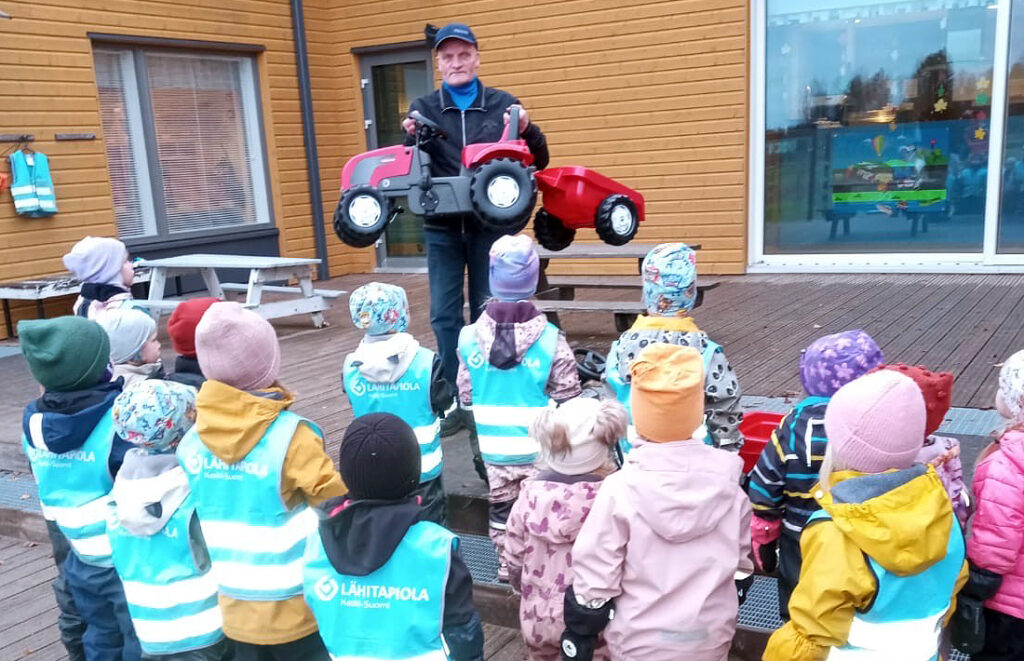 Lapsia kuvattuna takaapäin ulkona. Heidän edessään mies ojentamassa punaista polkutraktoria.