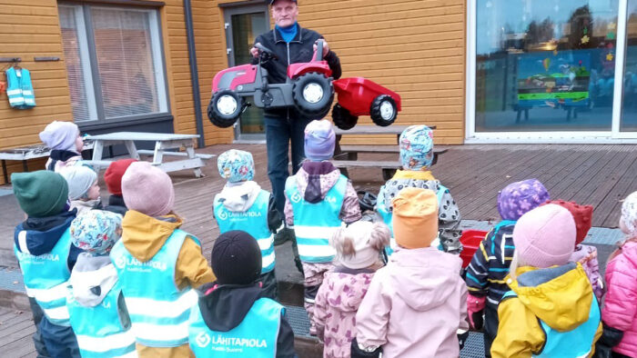 Lapsia kuvattuna takaapäin ulkona. Heidän edessään mies ojentamassa punaista polkutraktoria.