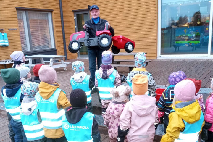 Lapsia kuvattuna takaapäin ulkona. Heidän edessään mies ojentamassa punaista polkutraktoria.