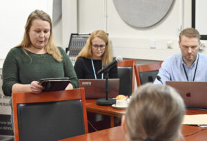 Naishenkilö jolla vihreä neule ja vaaleat pitkät hiukset seisoo pöydän takana käsissään tabletti, jota hän katsoo. 