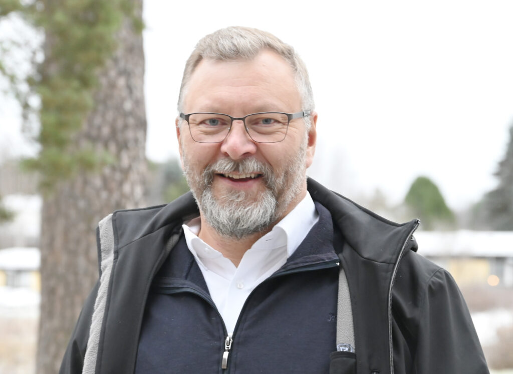 Mies seisomassa ulkona, kuvattuna puolilähikuvassa. Silmälasit, parta ja lyhyethiukset. Yllään kaulispaita ja musta takki.