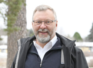 Mies seisomassa ulkona, kuvattuna puolilähikuvassa. Silmälasit, parta ja lyhyethiukset. Yllään kaulispaita ja musta takki.