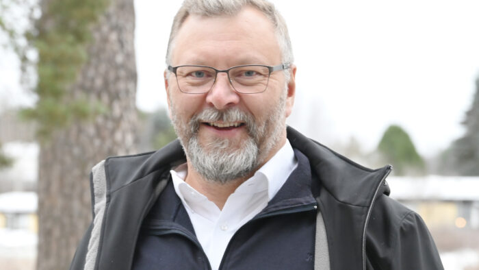 Mies seisomassa ulkona, kuvattuna puolilähikuvassa. Silmälasit, parta ja lyhyethiukset. Yllään kaulispaita ja musta takki.