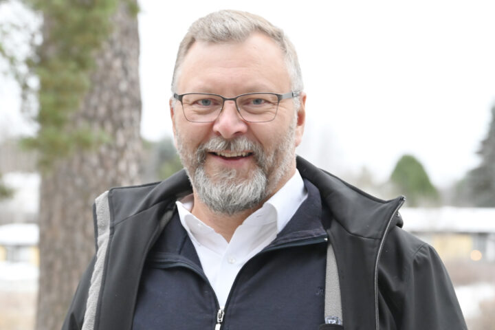 Mies seisomassa ulkona, kuvattuna puolilähikuvassa. Silmälasit, parta ja lyhyethiukset. Yllään kaulispaita ja musta takki.