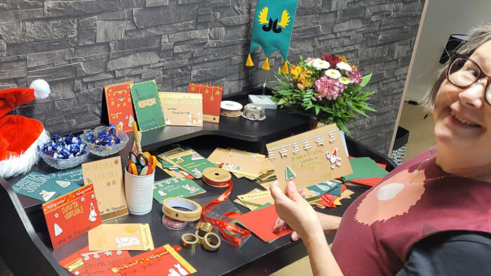 Pöytä jolla joulukortteja. Pöydän äärellä vasemmalla naishenkilö katsoo hymyillen kameraan