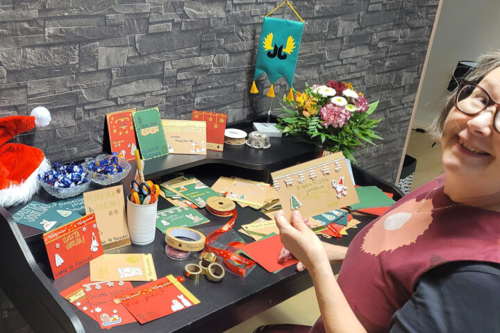 Pöytä jolla joulukortteja. Pöydän äärellä vasemmalla naishenkilö katsoo hymyillen kameraan