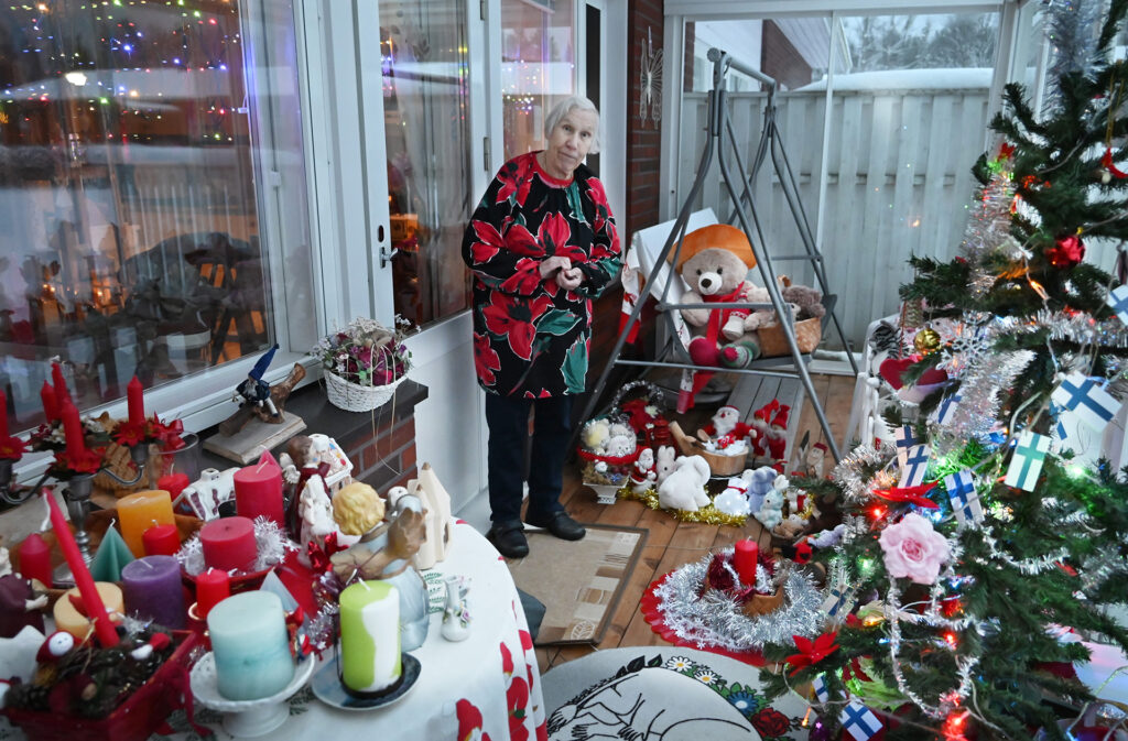 Terassi jolla leluja, kynttilöitä ja tonttuja. Myös joulukuusi. Keskellä seisoo naishenkilö, jolla harmaat hiukset kukallinen paita