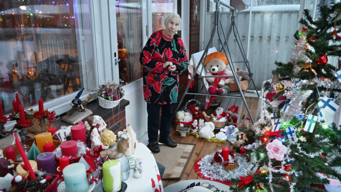 Terassi jolla leluja, kynttilöitä ja tonttuja. Myös joulukuusi. Keskellä seisoo naishenkilö, jolla harmaat hiukset kukallinen paita