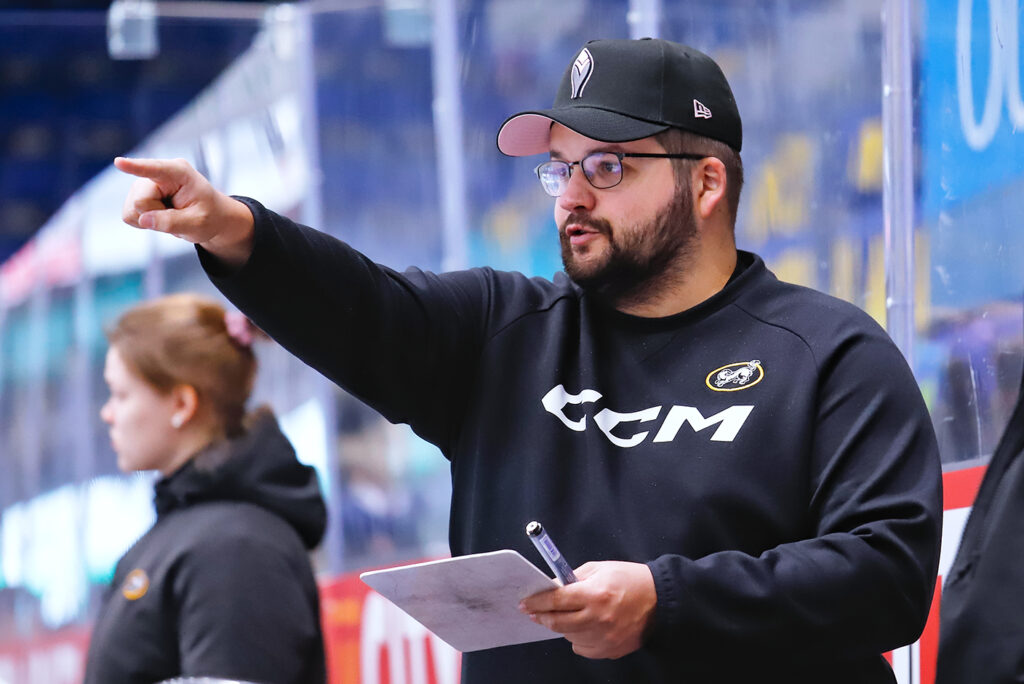 mieshenkilö, jolla lippalakki, silmälasit ja parta, puolilähikuvassa jääkiekkokaukalon laidalla. Hän katsoo sivulle ja osoittaa kädellään katseensa suuntaan.