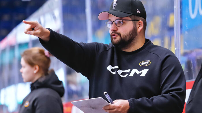 mieshenkilö, jolla lippalakki, silmälasit ja parta, puolilähikuvassa jääkiekkokaukalon laidalla. Hän katsoo sivulle ja osoittaa kädellään katseensa suuntaan.