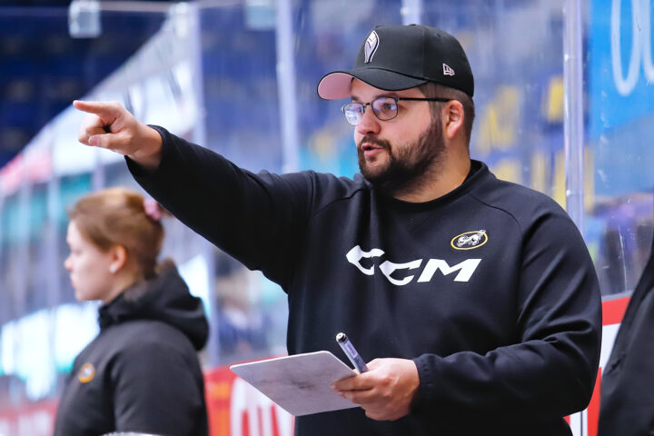 mieshenkilö, jolla lippalakki, silmälasit ja parta, puolilähikuvassa jääkiekkokaukalon laidalla. Hän katsoo sivulle ja osoittaa kädellään katseensa suuntaan.