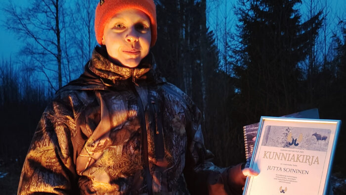 henkilö jolla yllään metsästysasu ja punainen pipo. Katsoo kameraan.