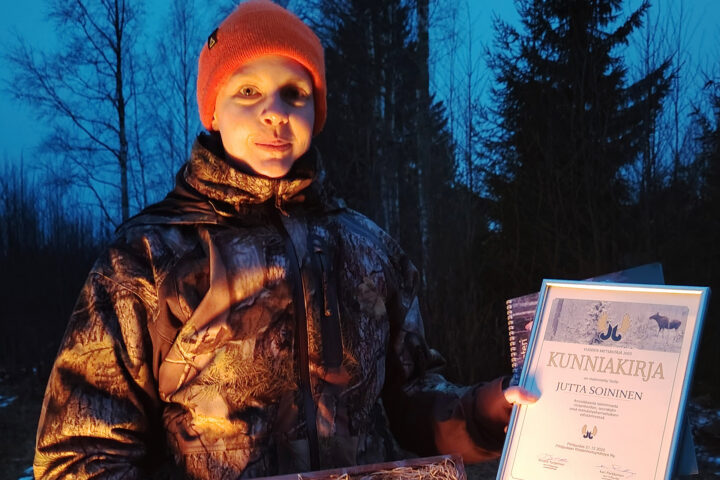 henkilö jolla yllään metsästysasu ja punainen pipo. Katsoo kameraan.