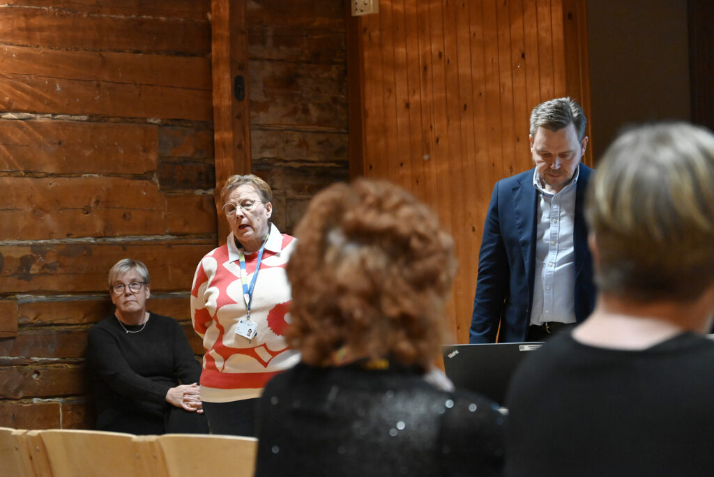 Kolme henkilöä, joista kaksi seisoo, yksi istuu. Katsotavat yleisöön, josta kuvassa näkyy kahden henkilön takaraivo kuvan etualalla.