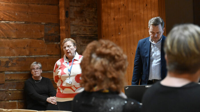 Kolme henkilöä, joista kaksi seisoo, yksi istuu. Katsotavat yleisöön, josta kuvassa näkyy kahden henkilön takaraivo kuvan etualalla.