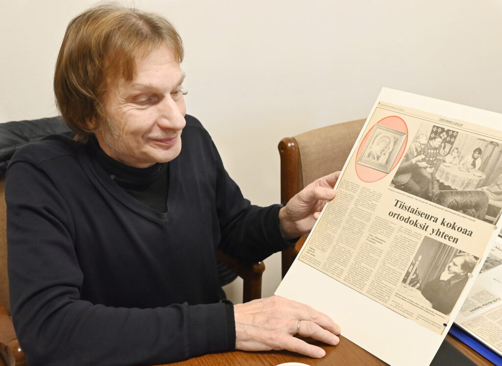 Mieshenkilö pöydän ääressä käsissään lehtileike. Puolikuva. miehellä on yllään musta paita.