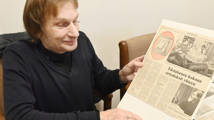 Mieshenkilö pöydän ääressä käsissään lehtileike. Puolikuva. miehellä on yllään musta paita.