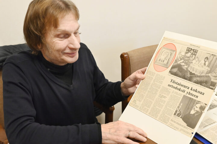 Mieshenkilö pöydän ääressä käsissään lehtileike. Puolikuva. miehellä on yllään musta paita.