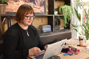 naishenkilö istuu tietokoneensa äärellä. Polkkatukka, silmälasit ja musta neulepaita.