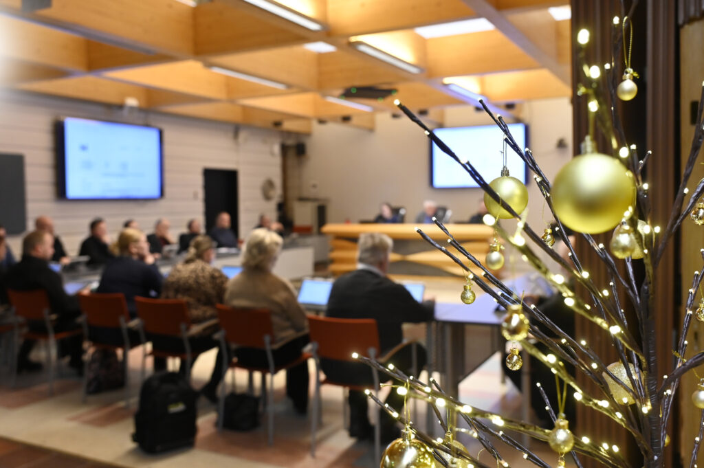 Huone, jossa istuu henkilöitä puoliympyrän muodossa. Etualalla kuvassa oksia, joissa joulukoristeita.